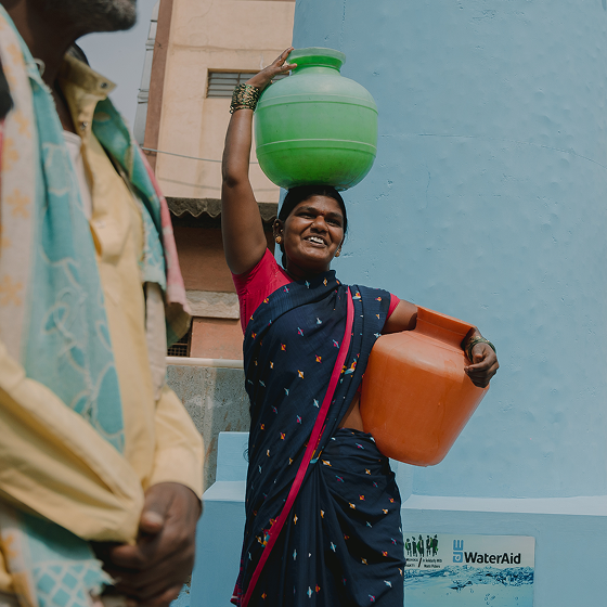 Woman carrying water containers on her head with a WaterAid logo in the background Woman carrying water containers on her head with a WaterAid logo in the background