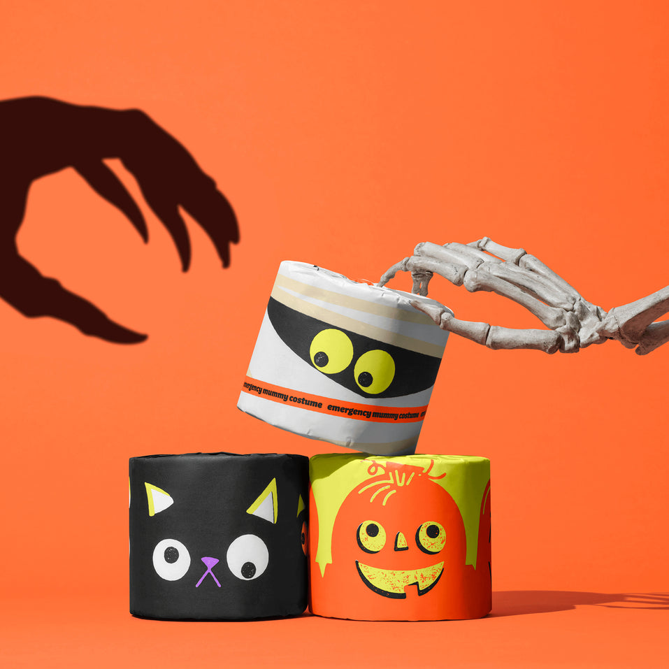 Skeleton hand holding mummy-wrapped Spooky Edition toilet paper with black cat and pumpkin rolls stacked below