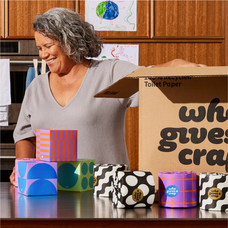 Person standing behind kitchen benchtop with toilet paper rolls and tissue boxes on the counter. Their hand reaches inside a Who Gives A Crap box.