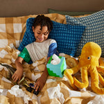 A smiling child is playing with toys in bed with multiple used tissues with a blue and green box of tissues nearby.