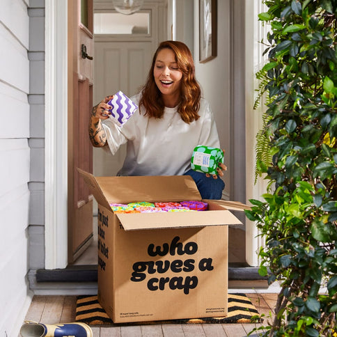 A woman unboxes a box of 48 Recycled Toilet Paper outside her door.