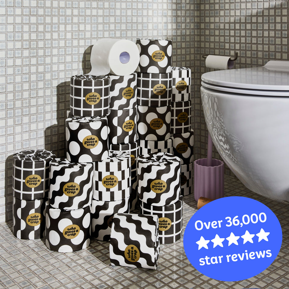 Several rolls of wrapped and unwrapped Premium Toilet Paper sits on the bathroom floor next to a toilet. Copy over the image reads ‘Over 36,000 5 star reviews’.