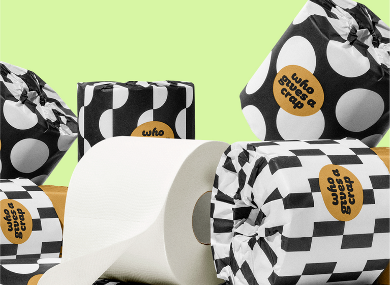A stack of Who Gives A Crap toilet paper rolls wrapped in black and white patterned paper, alongside a plain white unwrapped roll, set against a pale green background