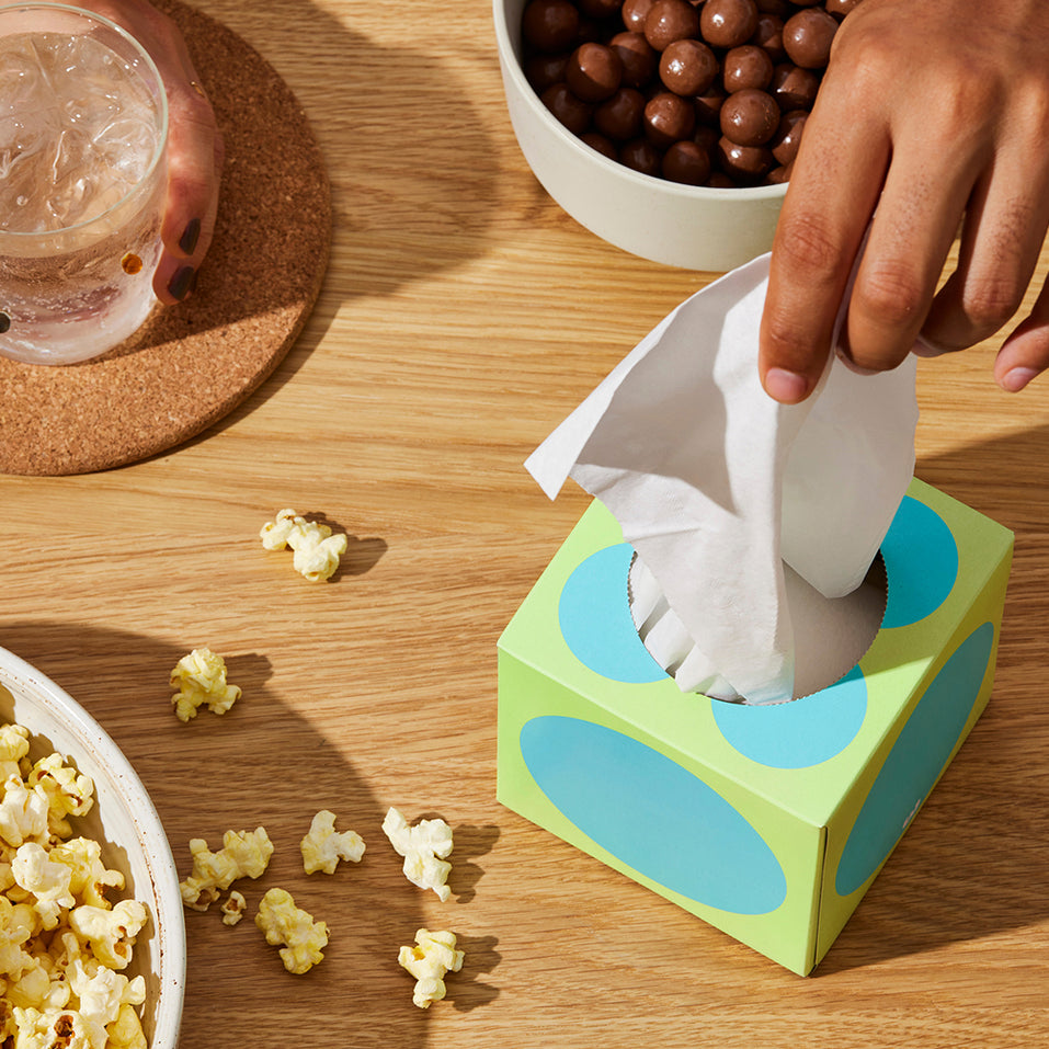 Hand grabs tissue from Who Gives a Crap tissue box on table with snacks and drinks