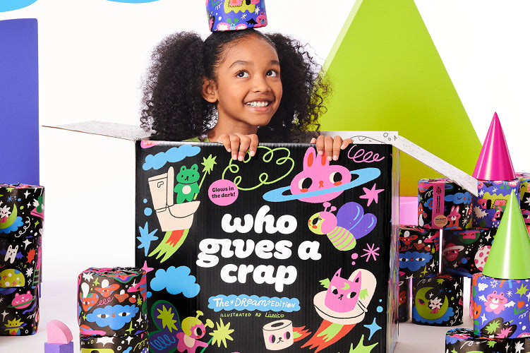 A child sitting inside a box of Dream Edition toilet paper surrounded by stacks of colourfully wrapped rolls and fun illustrative shapes and animals.