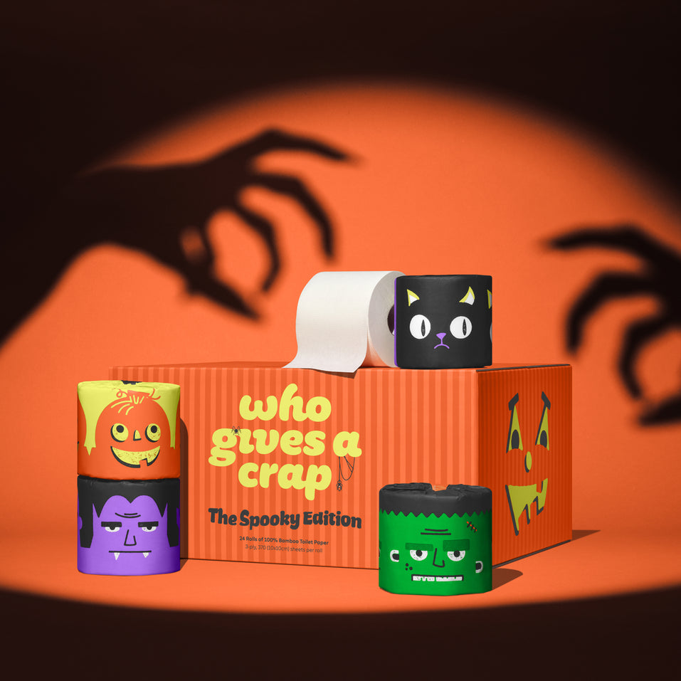 Who Gives A Crap Spooky Edition 100% Bamboo toilet paper set with Halloween-themed wrappers and designs against orange backdrop with creepy hand shadows
