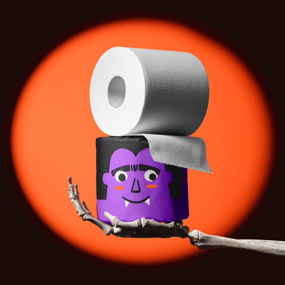 100% Bamboo toilet roll stacked on top of Dracula-themed Spooky Edition wrapper held by skeleton hand in front of orange spotlight backdrop