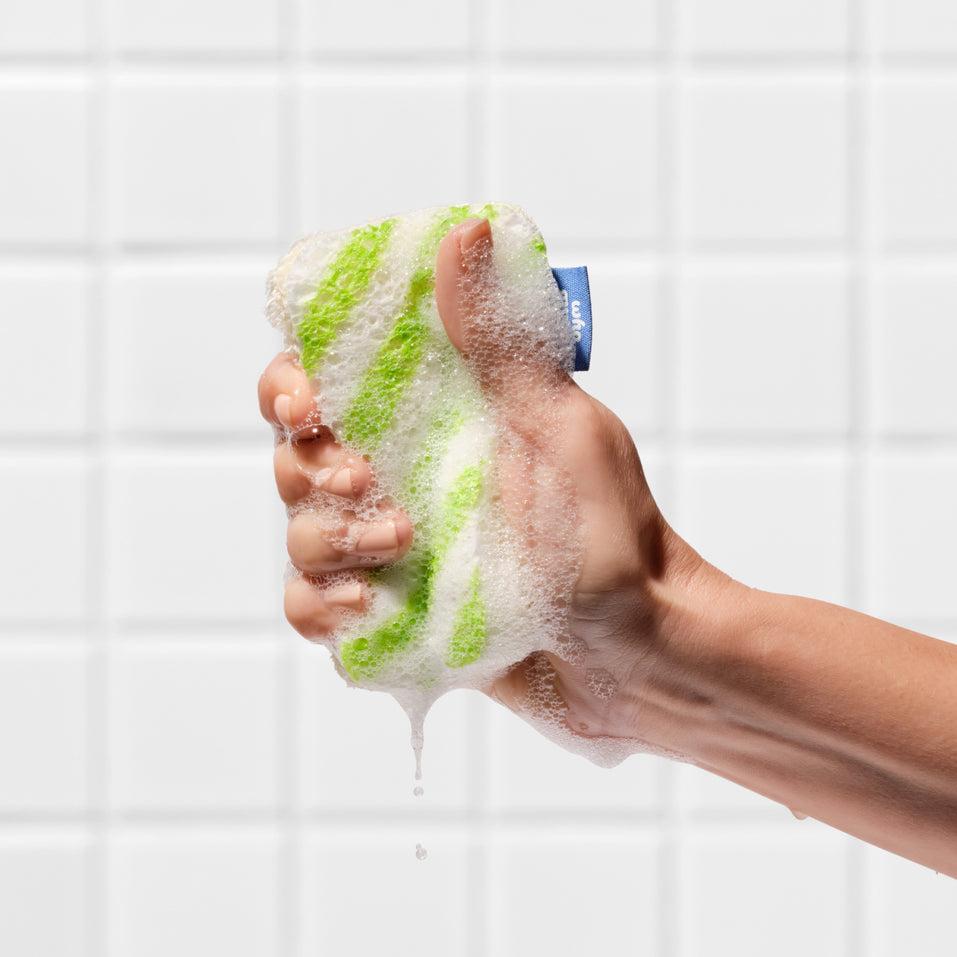 A hand squeezing a lathered Who Gives A Crap green striped scrub sponge against a tiled wall, with soapy water dripping down