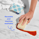 A hand scrubbing a surface with a Who Gives A Crap natural loofah scrub sponge next to a blue polka dot sponge, with a callout highlighting that the sponges are reusable and machine washable