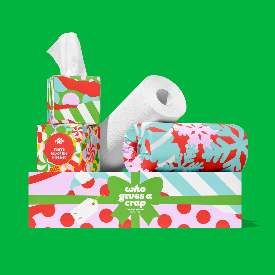 The Perfect Pair: Who Gives A Crap Gift Edition tissue boxes and wrapped paper towel rolls in festive red, pink, green and blue designs, stacked on a gift box on a green background.