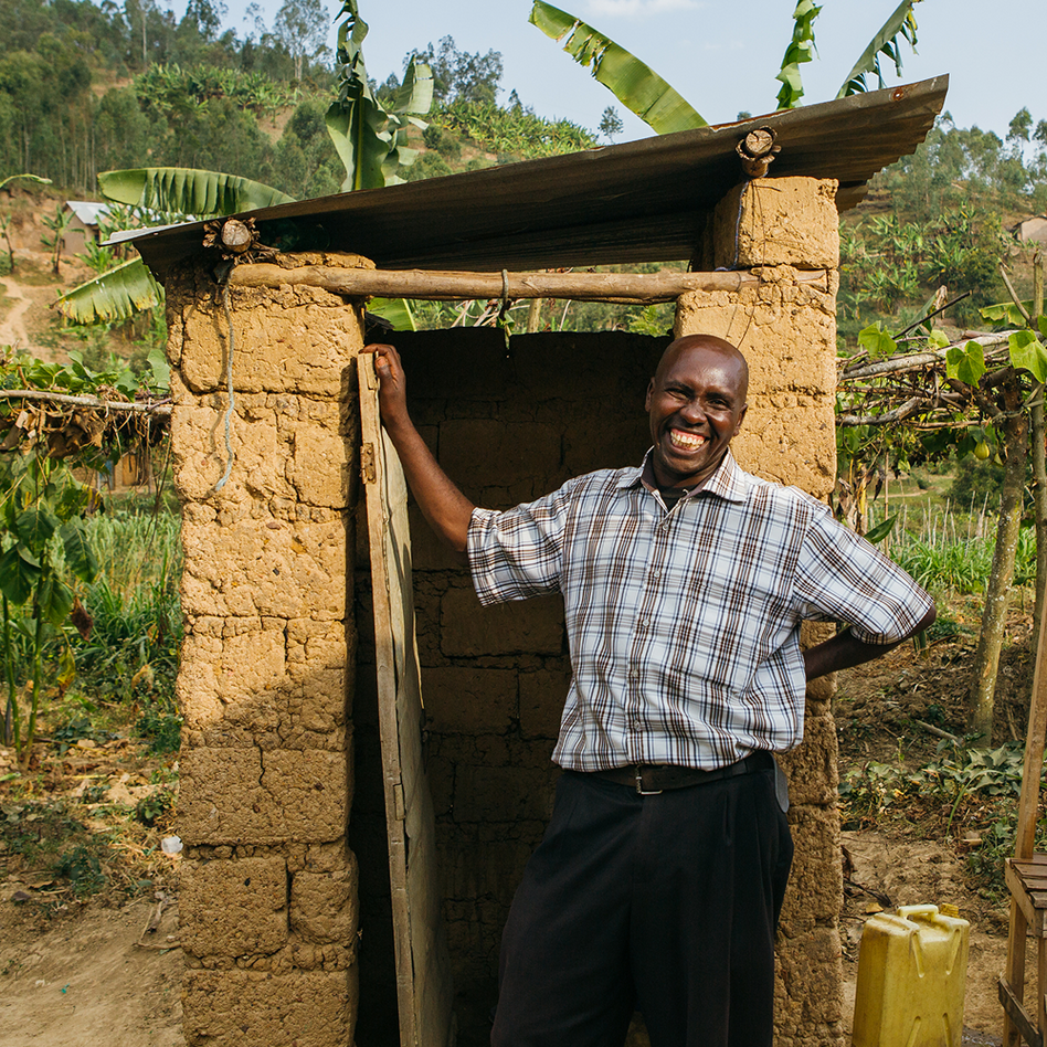 Man standing and smiling in the doorway of a brick toilet block in Rwanda, banana plants growing behind it