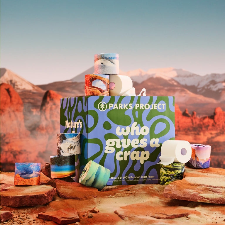 Who Gives A Crap x Parks Project product box and stacked national-park-design rolls on red-rock desert terrain with distant buttes and a pale sky.