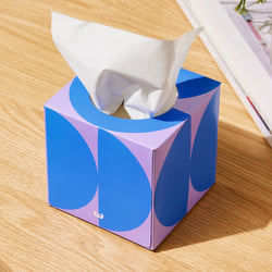 A square tissue box with bold blue and purple geometric circle pattern, with a white tissue pulled out of the top, sitting on a wooden table.